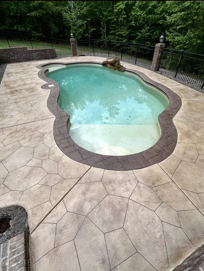 Stamped Concrete Pool Deck by Alex's Lawnscape in Charlotte NC — Freeform pool surrounded by stamped concrete decking with decorative brick border, brick columns, and iron fencing in a wooded setting