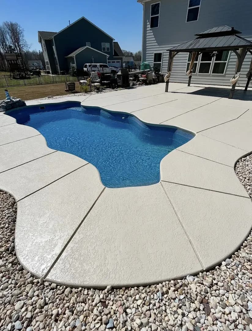 Pool & Concrete Surround by Alex's Lawnscape in Charlotte NC — Freeform fiberglass pool with clean poured concrete surround, river rock border, and gazebo in a residential backyard