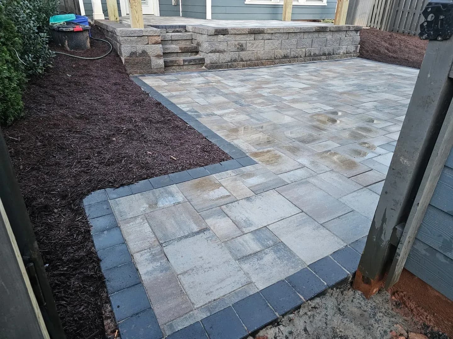 Two-Tier Patio with Accent Border by Alex's Lawnscape in Charlotte NC — Multi-level paver patio with dark border edging, retaining wall block steps, and fresh mulch beds at a blue-sided home