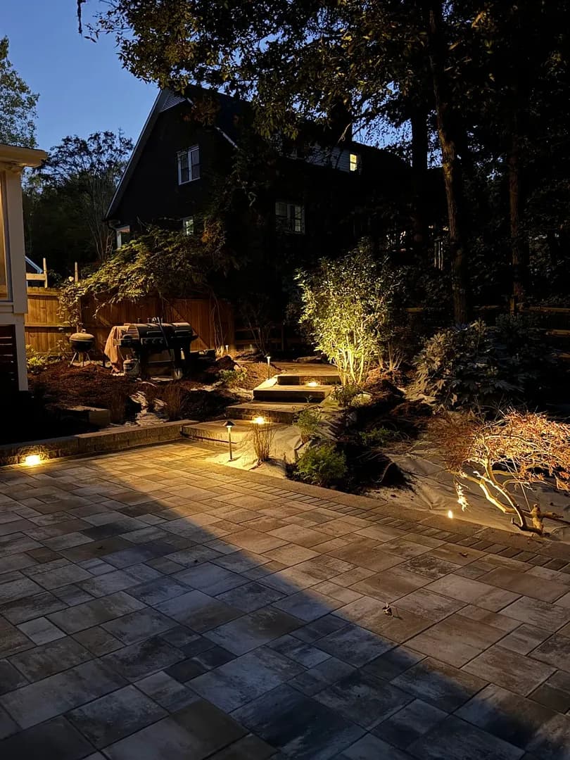 Artificial Turf Backyard by Alex's Lawnscape in Charlotte NC — Lush synthetic turf lawn edged with river rock and pavers, retaining wall with step lights, in a shaded backyard