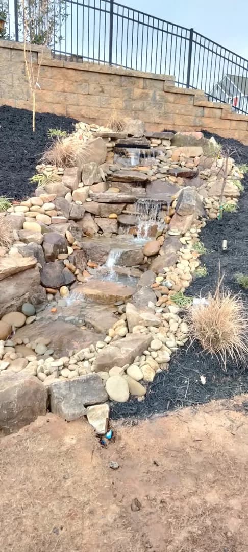 Stone Waterfall at Brick Building by Alex's Lawnscape in Charlotte NC — Natural stone waterfall feature with river stones and flagstone path edge adjacent to a brick building