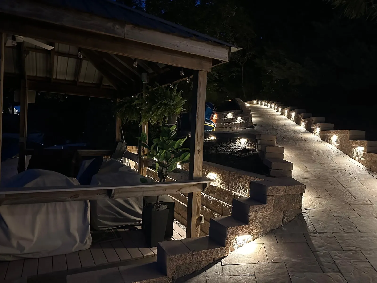 Pavilion & Landscape Lighting by Alex's Lawnscape in Charlotte NC — Covered pavilion with timber framing, multi-tier stone retaining walls with integrated LED lighting, paver walkway, and hanging fern planters