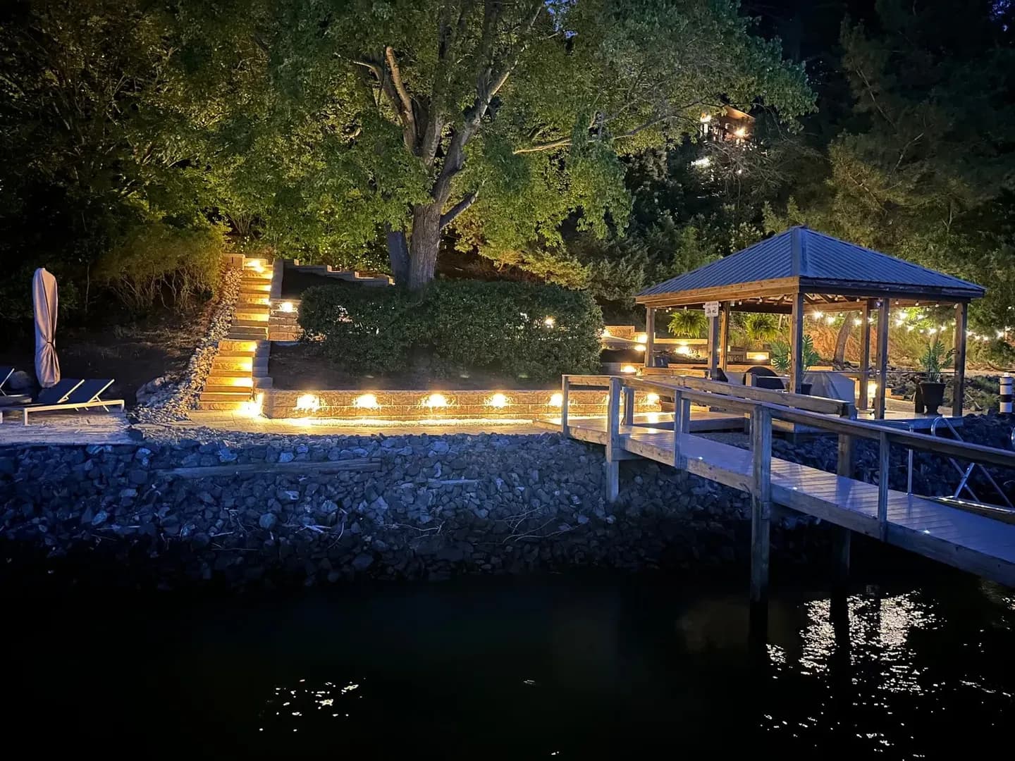 Lakefront Outdoor Living Space by Alex's Lawnscape in Charlotte NC — Full lakefront build — tiered stone retaining walls, built-in stone staircase with step lighting, covered gazebo with string lights, and riprap shoreline stabilization