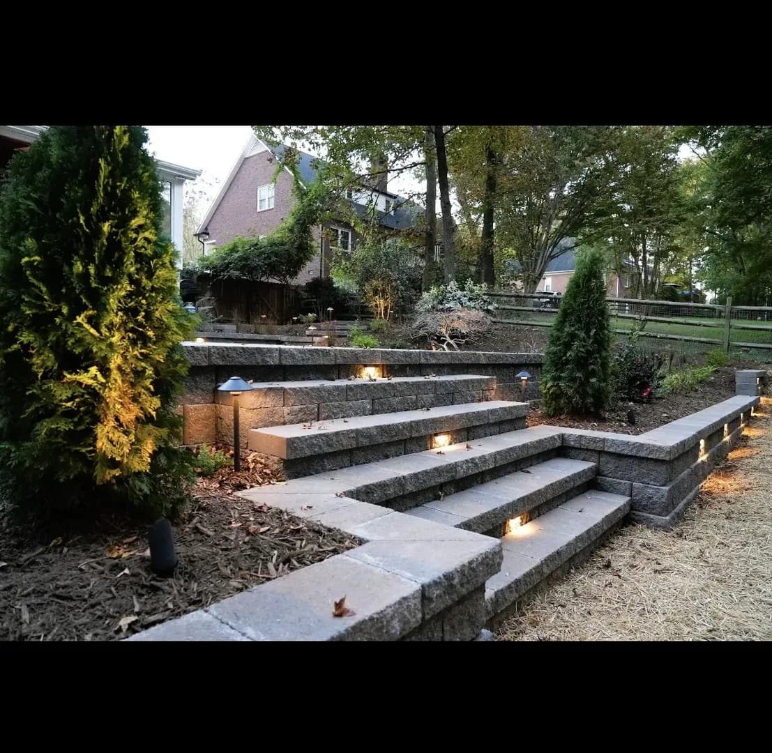 Stone Retaining Wall Steps with Lighting by Alex's Lawnscape in Charlotte NC — Multi-level stone block retaining wall with built-in staircase, recessed LED step lights, path lighting, and evergreen plantings along both sides