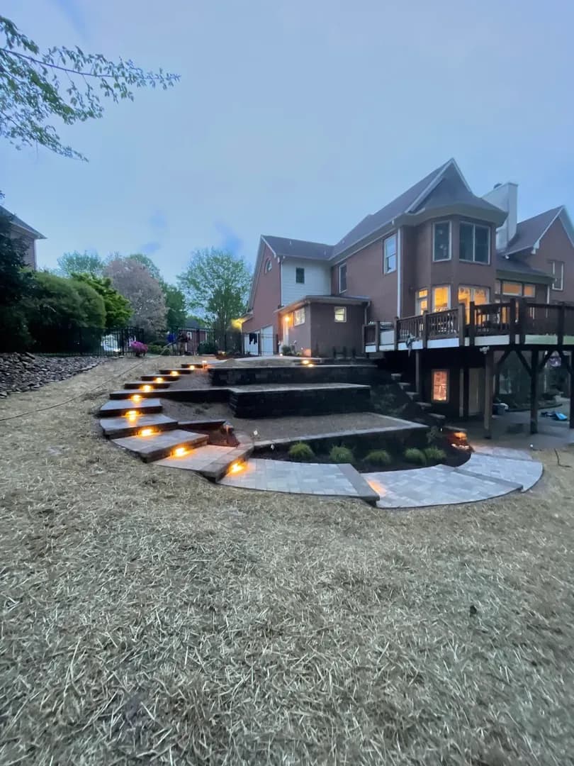 Tiered Paver Steps with LED Lighting by Alex's Lawnscape in Charlotte NC — Multi-level paver staircase with integrated LED riser lights cascading down a sloped backyard to a curved paver patio with fresh plantings and landscape lighting